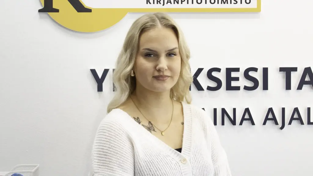 Siiri Montonen kuvassa valkoisen seinän edessä, jossa Kirjanpitotoimisto Kalevan logo ja slogan "Yrityksesi talous – aina ajallaan ja oikein".